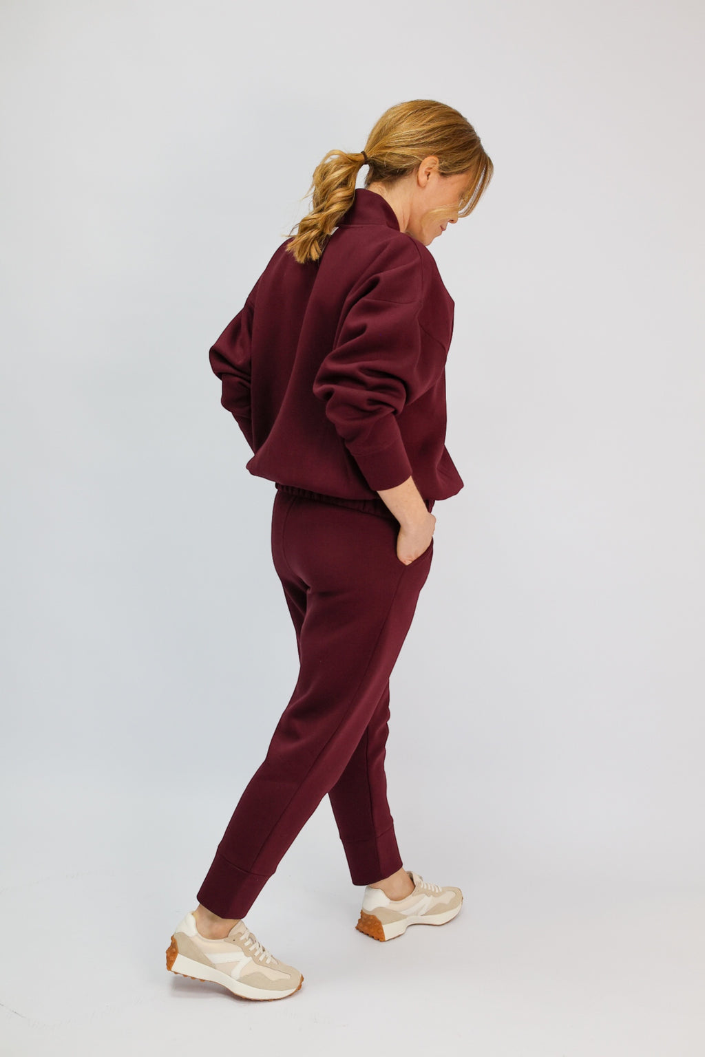 A woman wearing a matching burgundy sweatshirt and jogger pants set, shown from a side-back angle against a light gray studio background.