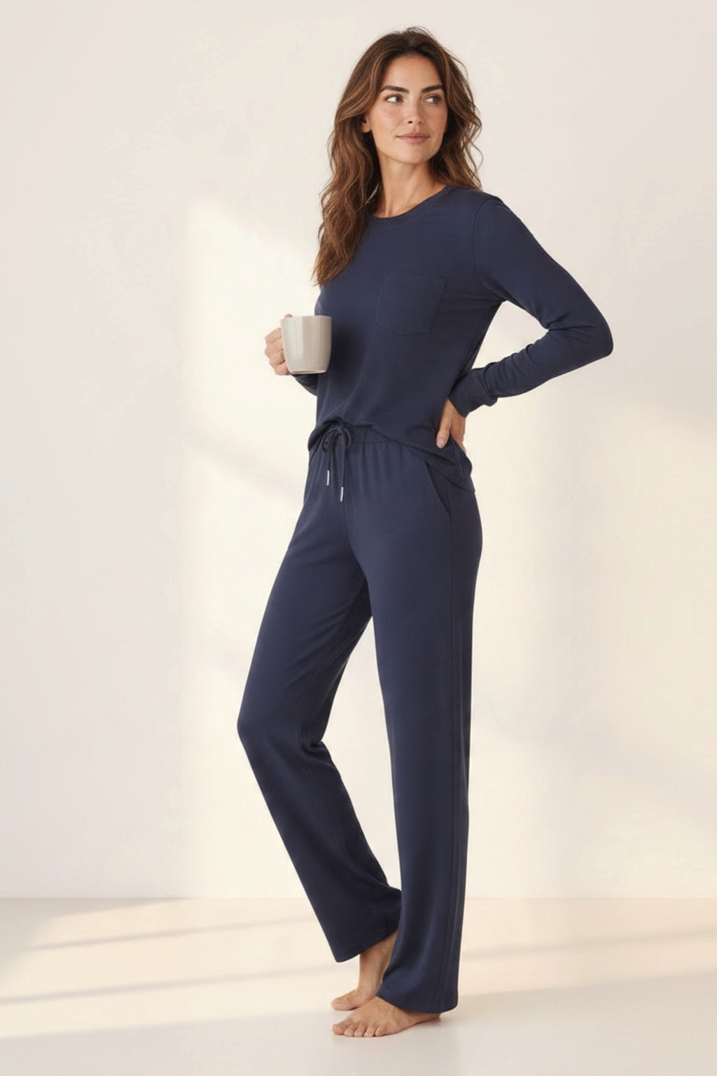 A woman modeling a navy blue long-sleeve lounge set with matching drawstring pants, standing barefoot in a bright studio while holding a white mug.