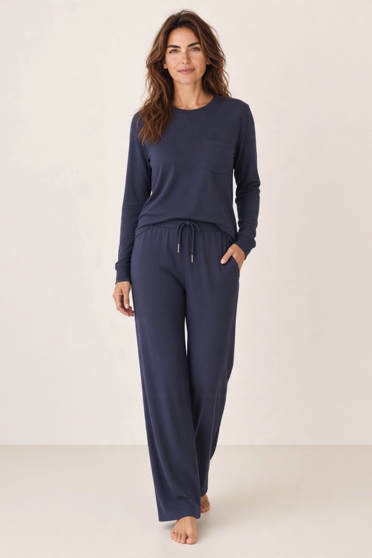 A woman modeling a matching navy blue lounge set consisting of a long-sleeve top with a chest pocket and matching drawstring wide leg pants.