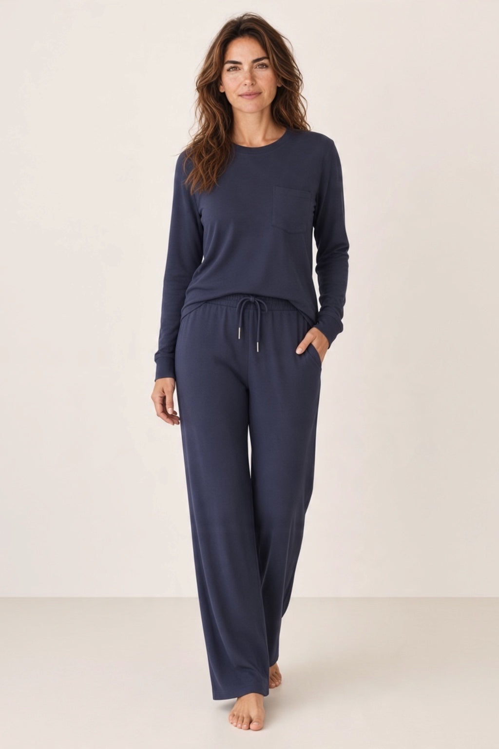 A woman modeling a matching navy blue lounge set consisting of a long-sleeve top with a chest pocket and matching drawstring pants.