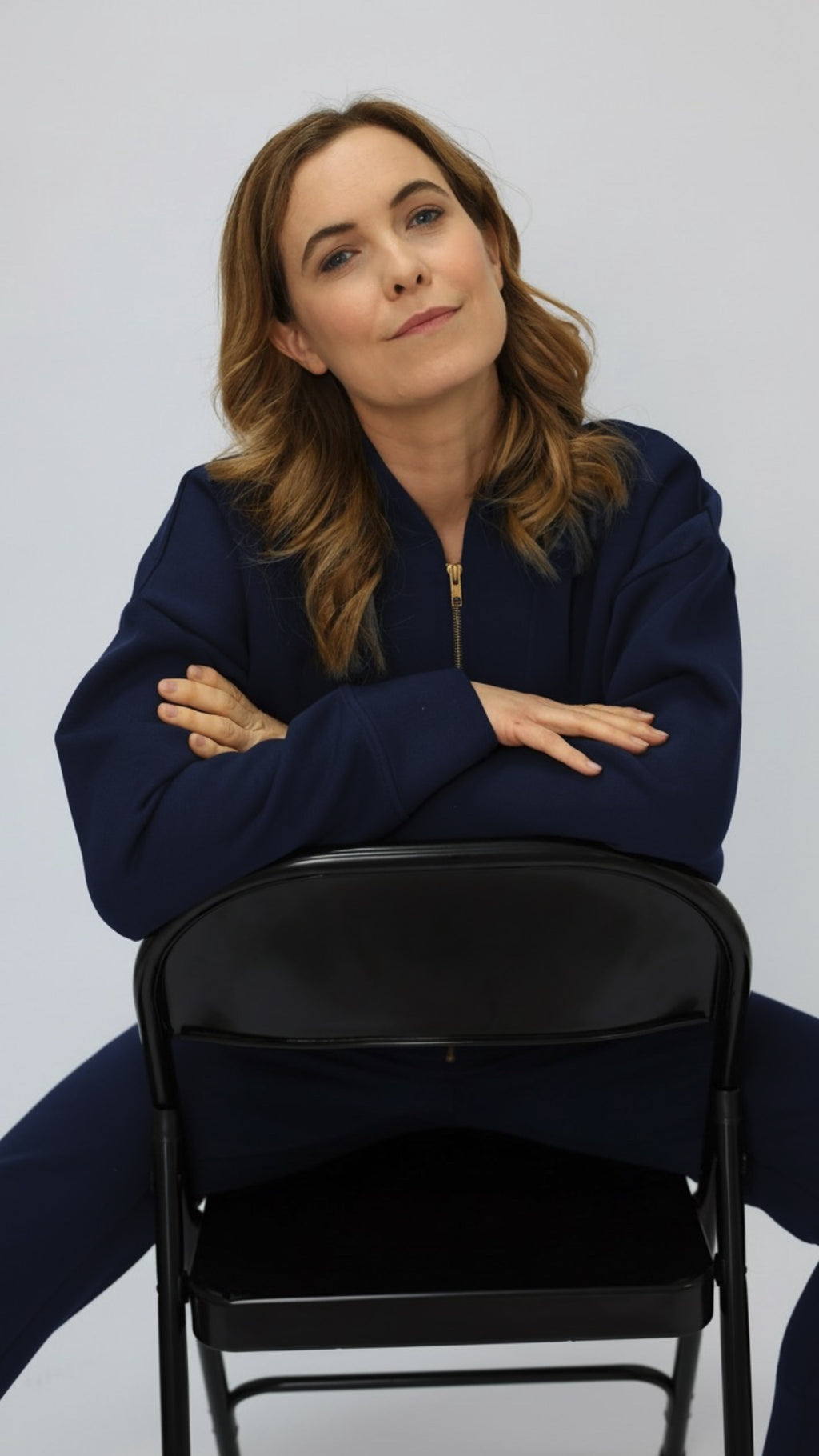 Woman in a navy blue sweatshirt and sweatpants sitting on a black chair against a plain background