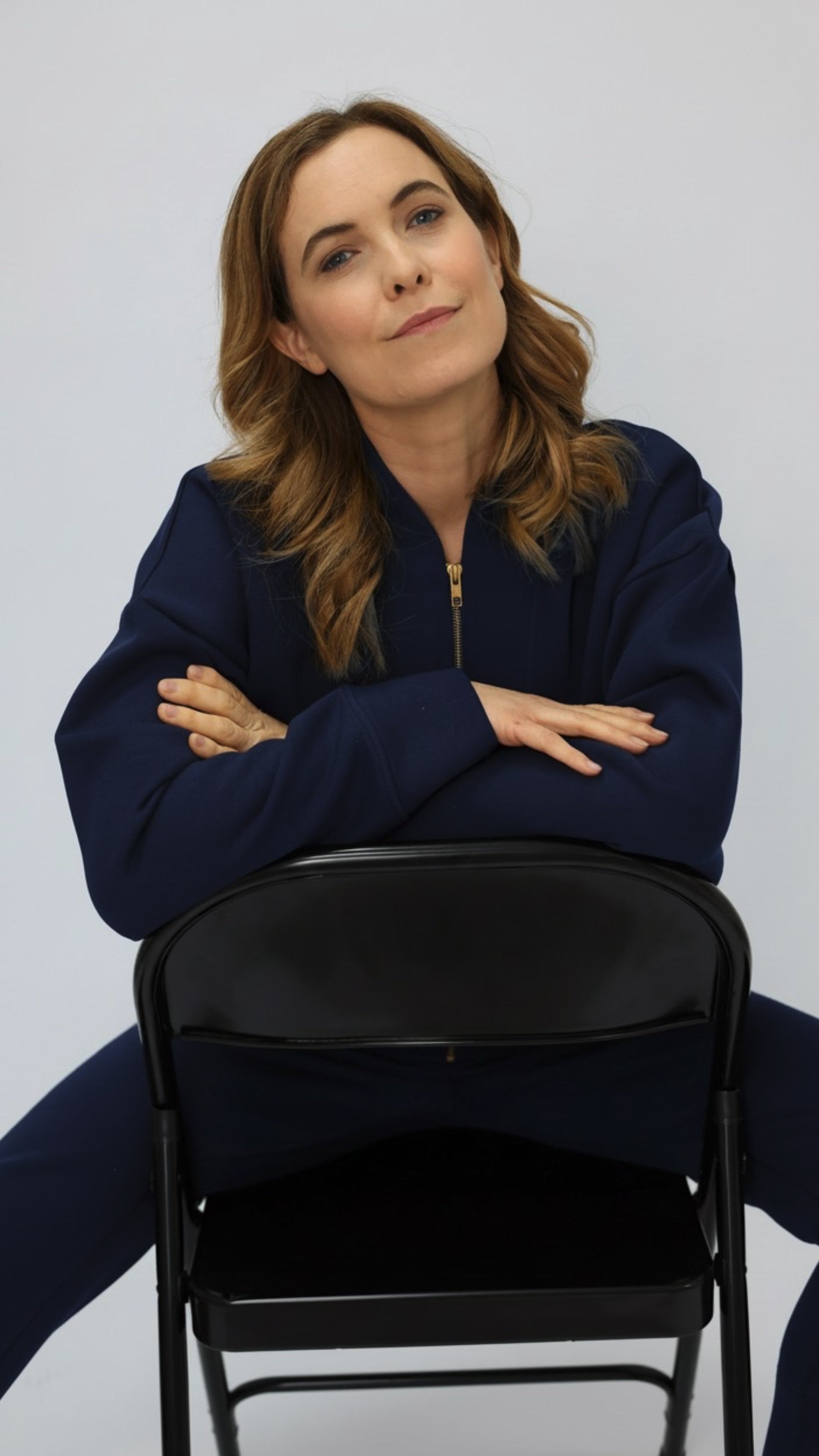 Woman in a navy blue sweatshirt and sweatpants sitting on a black chair against a plain background