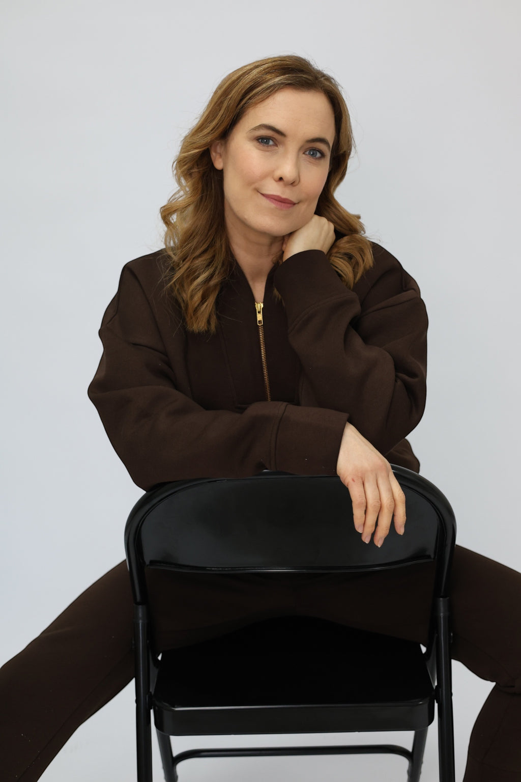 A woman with light brown hair sits on a black folding chair, wearing a dark brown zip-up jacket and matching brown pants against a light gray studio background.