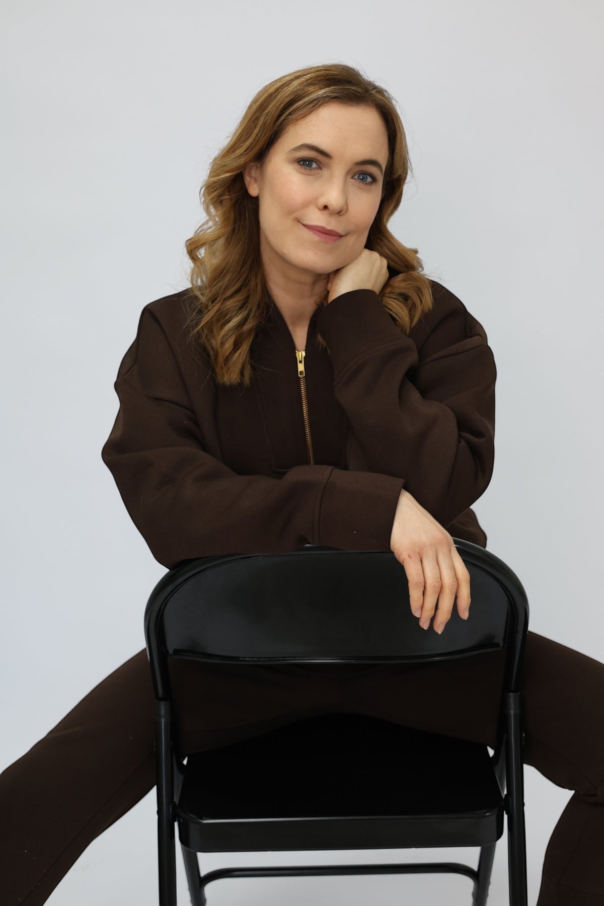 A woman with light brown hair sits on a black folding chair, wearing a dark brown zip-up jacket and matching brown pants against a light gray studio background.