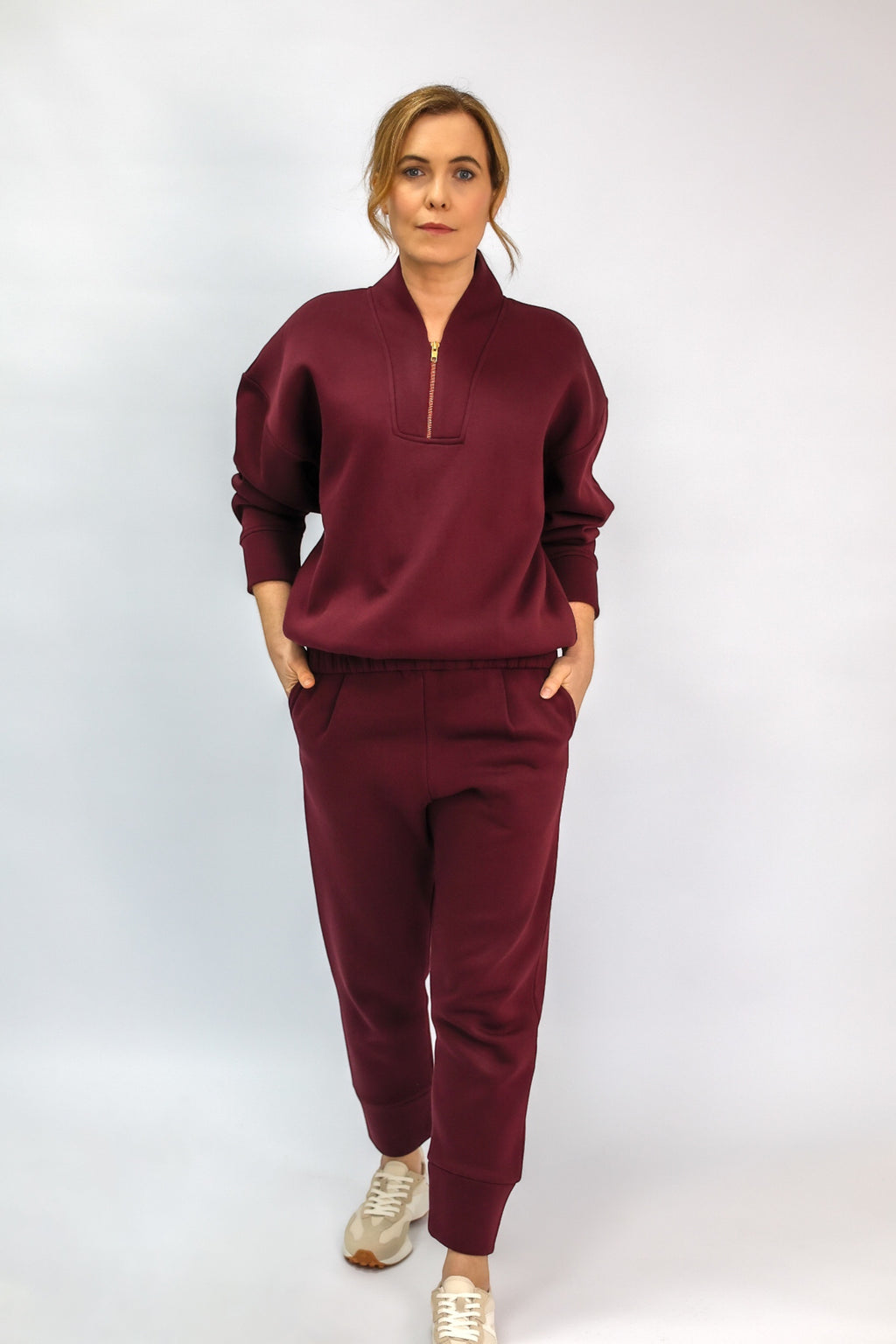 A woman modeling a matching burgundy tracksuit, featuring a half-zip sweatshirt and matching jogger pants, against a light gray studio background.