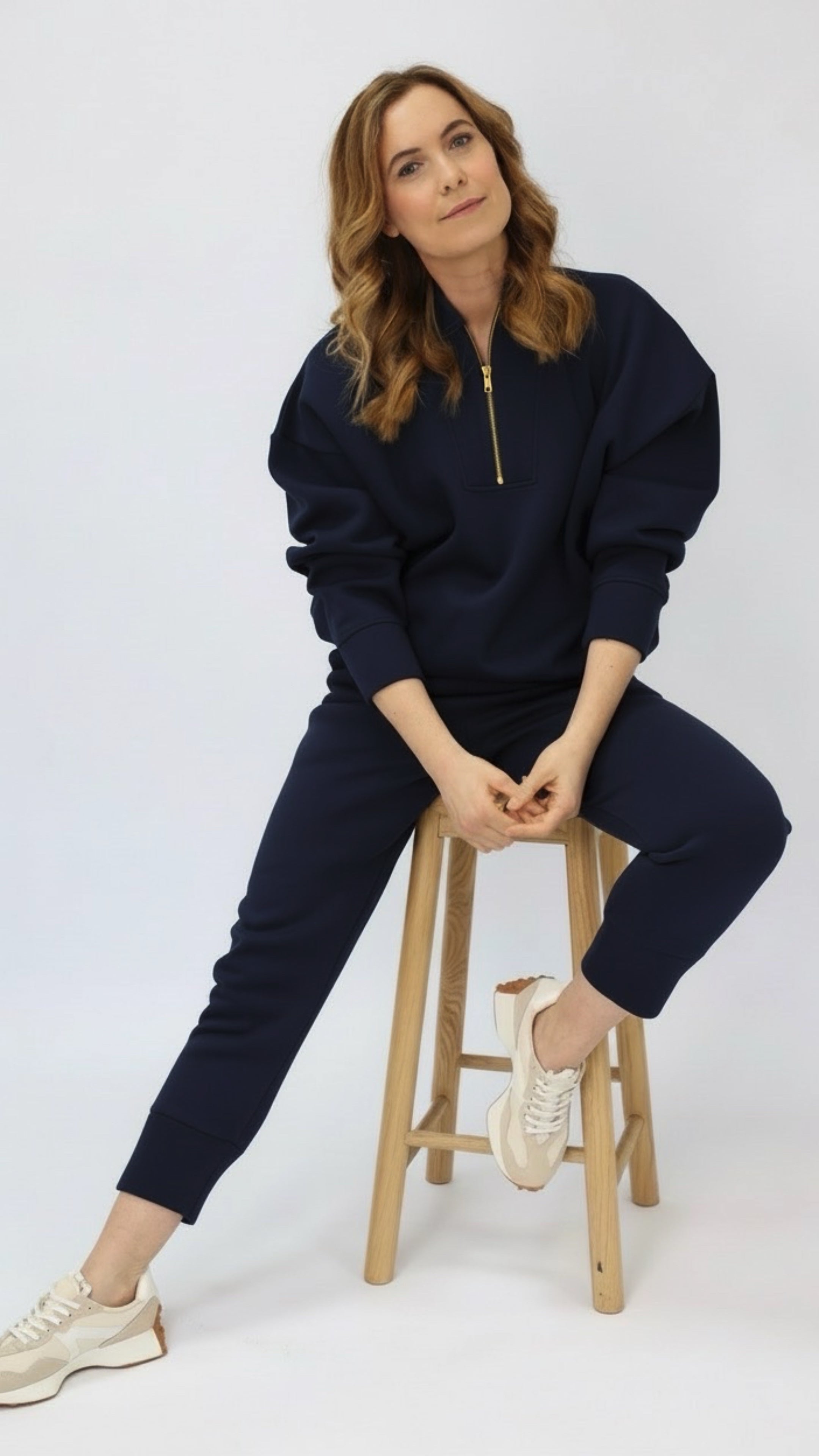 Woman wearing a navy blue tracksuit sitting on a wooden stool against a white background
