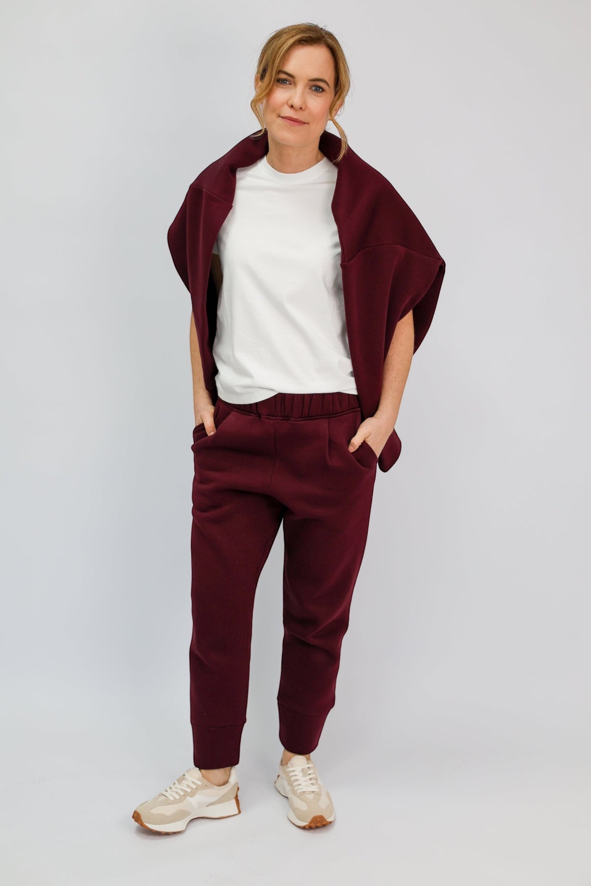 A woman modeling a matching burgundy lounge set consisting of jogger pants and a draped cardigan, styled with a white t-shirt and beige sneakers against a light gray studio background.