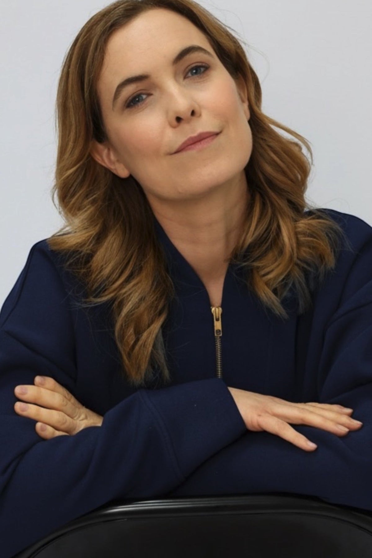 A woman with shoulder-length wavy brown hair wearing a navy blue half-zip sweatshirt, posing with her arms crossed.