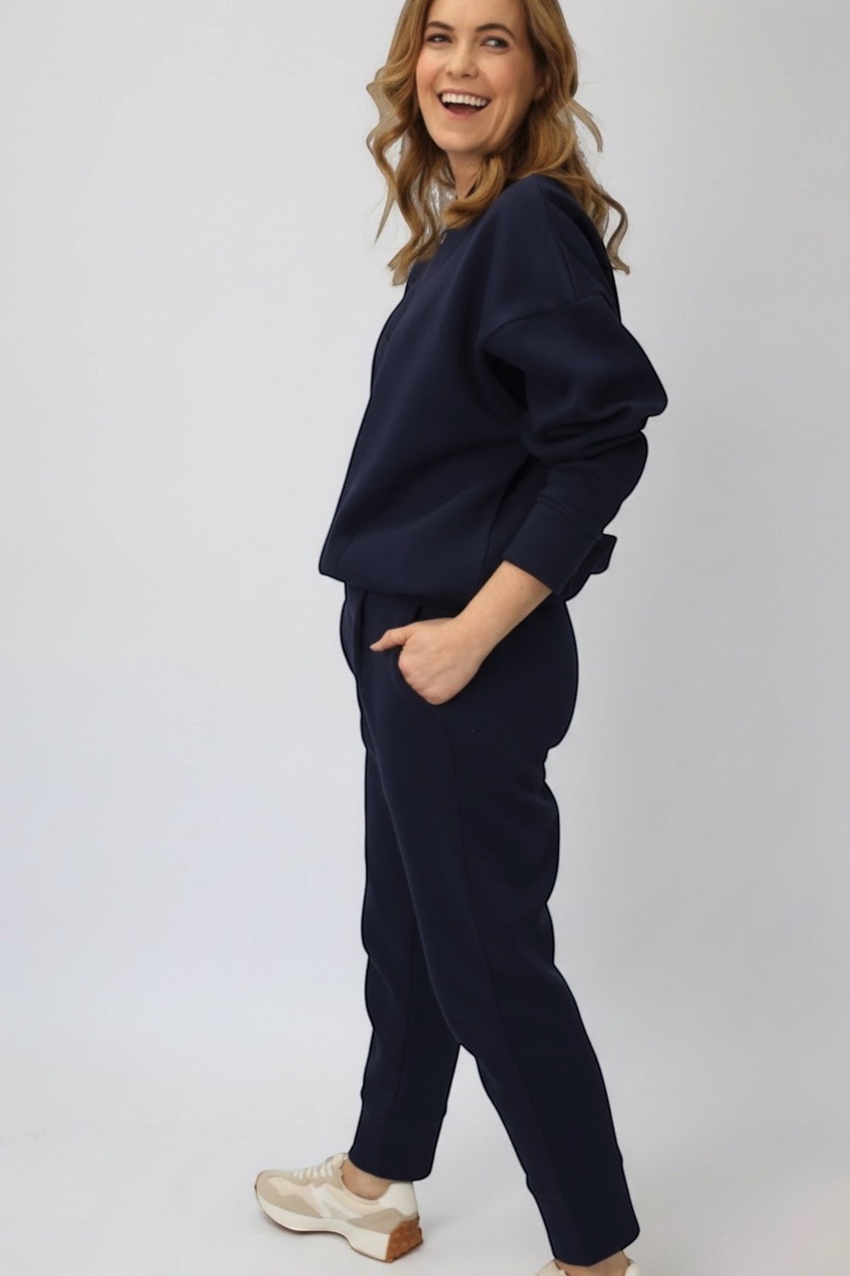 A woman modeling a matching navy blue lounge set consisting of a long-sleeve sweatshirt and jogger pants, paired with beige sneakers, against a plain light gray background.