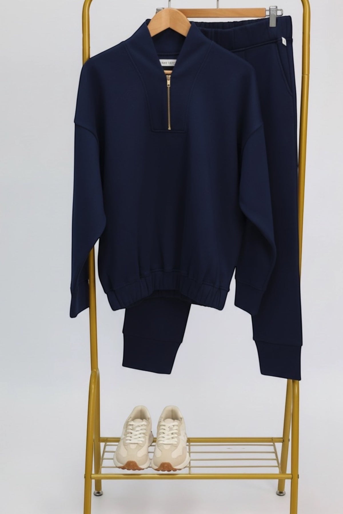 A navy blue two-piece tracksuit consisting of a quarter-zip pullover sweatshirt and matching pants, displayed on a gold clothing rack with a pair of cream sneakers on the shelf below.